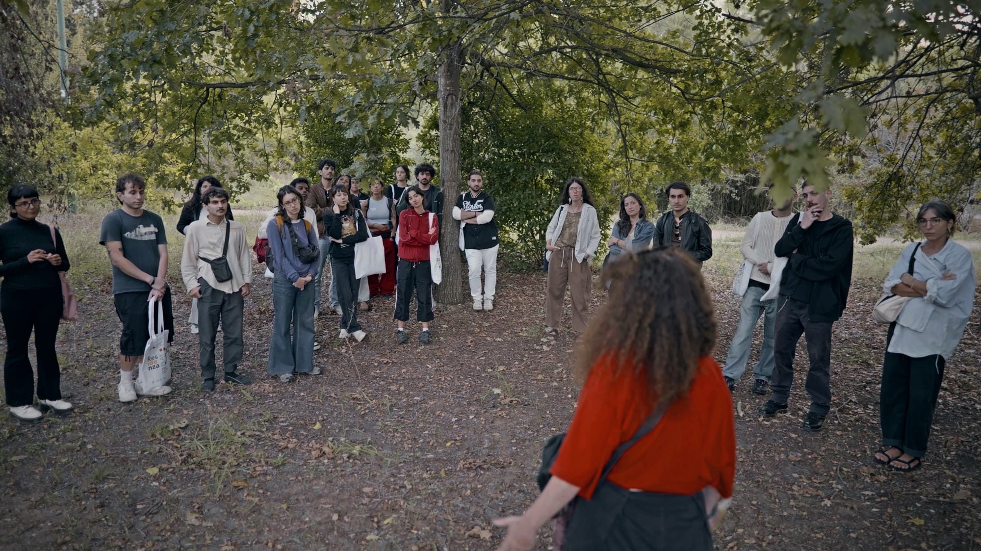 Una ventina di giovani in semicerchio sotto gli alberi ascolta una donna che parla, di spalle in primo piano.